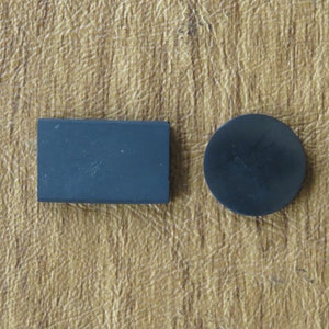 Puede incluir: Dos objetos pequeños de color azul oscuro sobre una superficie texturizada y marrón claro. Uno es rectangular, el otro es un círculo pequeño. Los objetos parecen estar hechos de un material sólido, posiblemente piedra o metal.