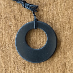 Könnte beinhalten: Ein schwarzer runder Anhänger mit einem Loch in der Mitte hängt an einer schwarzen Schnur. Der Anhänger ist aus einem glatten, polierten Material gefertigt.