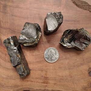 May include: Four pieces of black obsidian with iridescent sheen on a wooden surface. A US quarter is included for scale.