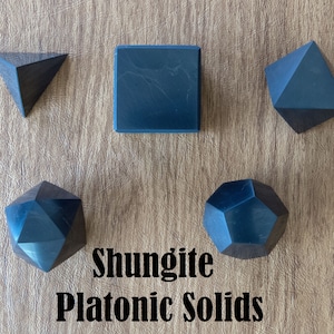 May include: Five black shungite platonic solids on a wooden surface. The solids are a cube, a tetrahedron, a dodecahedron, an octahedron, and an icosahedron. The text "Shungite Platonic Solids" is written below the solids.