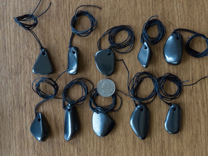 May include: A collection of dark gray, polished stone pendants on black cords. The pendants vary in shape, including teardrop, oval, and triangular designs. A US quarter is included for size reference. The pendants are displayed on a brown wooden surface.