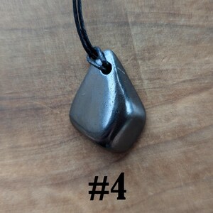 May include: A dark gray, polished stone pendant with a black cord necklace. The pendant is a smooth, rounded shape with a hole for the cord. The number "#4" is printed in black below the pendant.