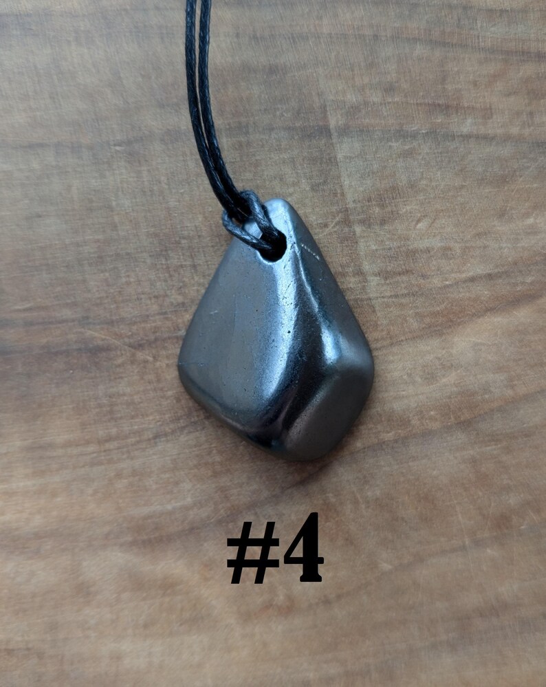 May include: A dark gray, polished stone pendant with a black cord necklace. The pendant is a smooth, rounded shape with a hole for the cord. The number "#4" is printed in black below the pendant.