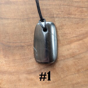 May include: A polished, dark gray pendant necklace with a rectangular shape. The pendant is strung on a black cord. The number "#1" is printed in black at the bottom of the image. The pendant is approximately 2 inches long.