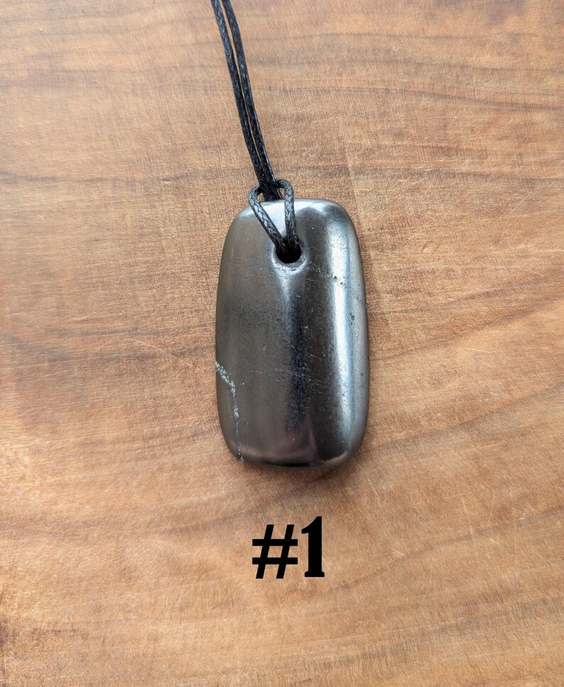 May include: A polished, dark gray pendant necklace with a rectangular shape. The pendant is strung on a black cord. The number "#1" is printed in black at the bottom of the image. The pendant is approximately 2 inches long.