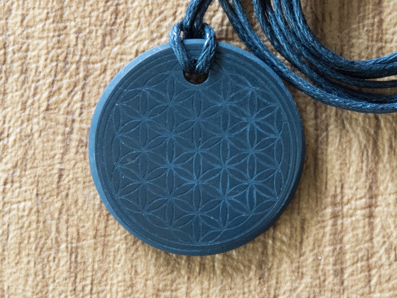 May include: A black pendant with a carved flower of life design, hanging on a black cord.