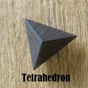 May include: A dark brown wooden tetrahedron, a geometric shape with four triangular faces. The tetrahedron is resting on a light brown surface.