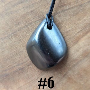 May include: A polished, dark gray gemstone pendant with a smooth, rounded shape, suspended from a black cord. The pendant is displayed against a wooden background, with the number "#6" in black text below it.