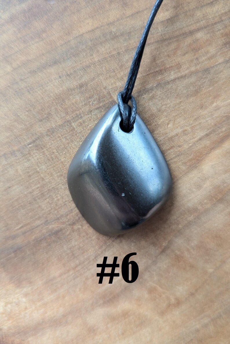 May include: A polished, dark gray gemstone pendant with a smooth, rounded shape, suspended from a black cord. The pendant is displayed against a wooden background, with the number "#6" in black text below it.