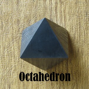 May include: A black octahedron, a geometric shape with eight triangular faces, is shown on a textured, light brown surface. The word "Octahedron" is printed in black below the shape.