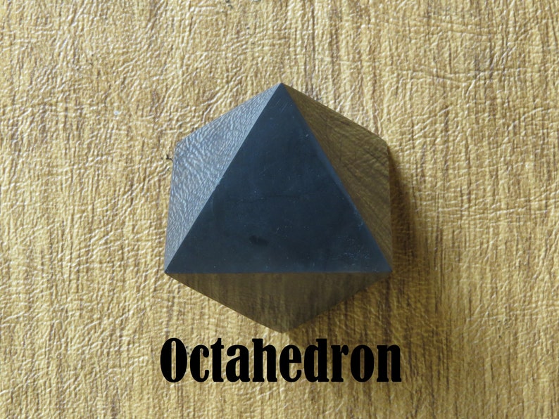 May include: A black octahedron, a geometric shape with eight triangular faces, is shown on a textured, light brown surface. The word "Octahedron" is printed in black below the shape.