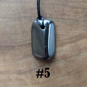 May include: A polished, rectangular, dark gray pendant with rounded edges, suspended from a black cord. The pendant has a smooth, reflective surface. The number "#5" is printed in black below the pendant. The background is a wooden surface.