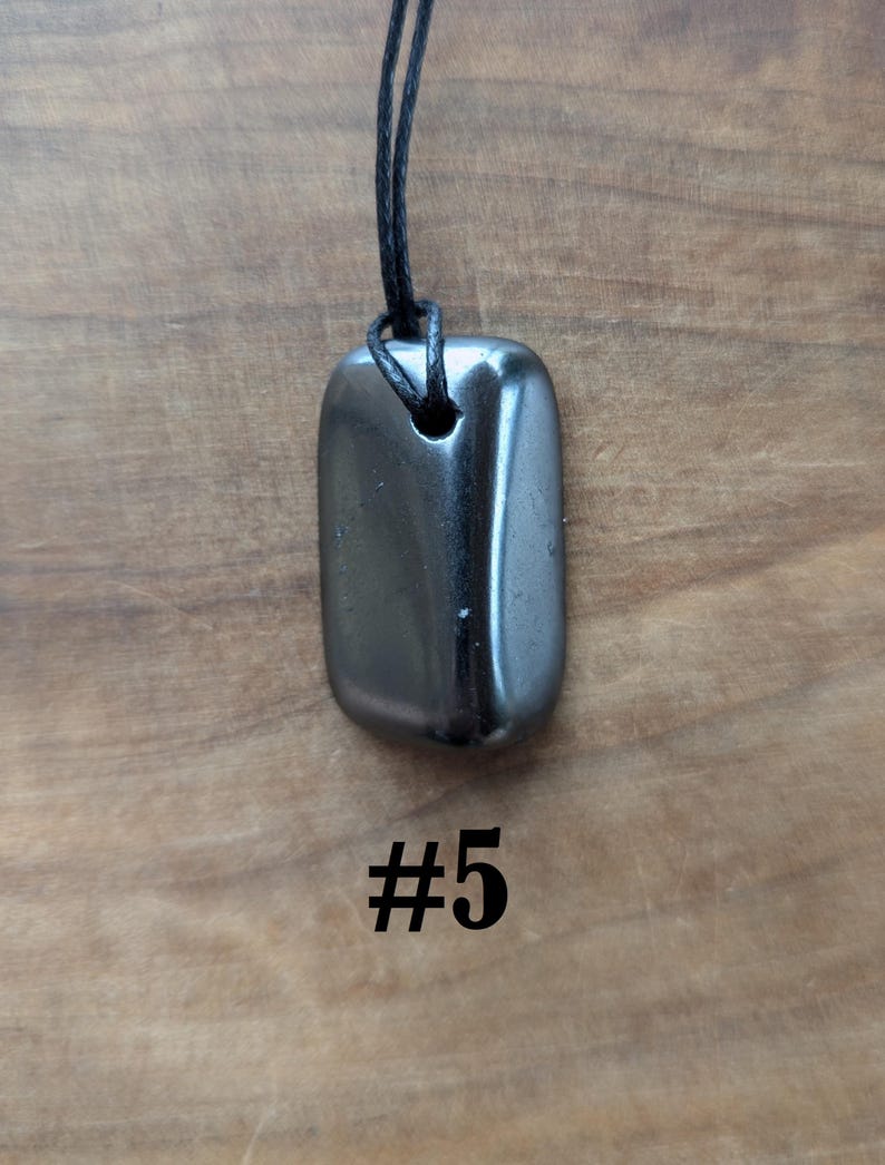 May include: A polished, rectangular, dark gray pendant with rounded edges, suspended from a black cord. The pendant has a smooth, reflective surface. The number "#5" is printed in black below the pendant. The background is a wooden surface.