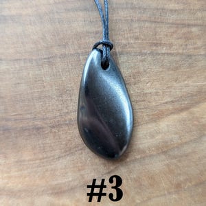 May include: A dark, teardrop-shaped pendant necklace with a black cord. The pendant is a polished, metallic gray stone with subtle swirling patterns. The number "#3" is printed in black at the bottom of the image. The necklace is displayed on a wooden surface.