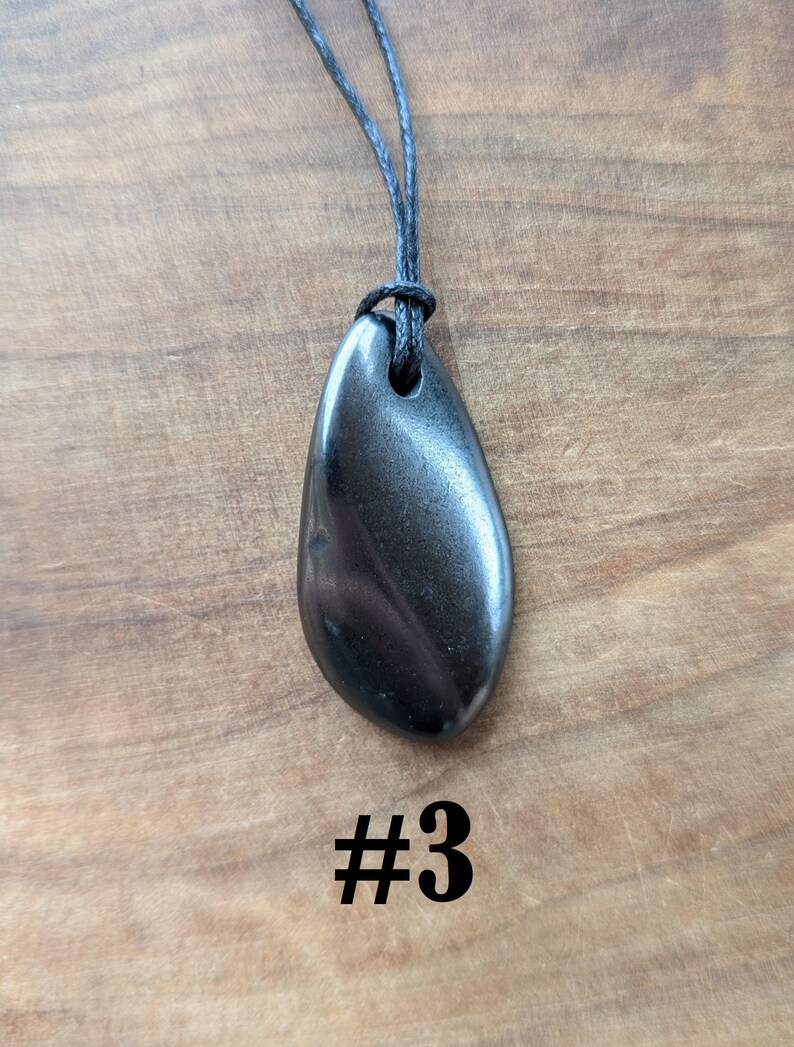 May include: A dark, teardrop-shaped pendant necklace with a black cord. The pendant is a polished, metallic gray stone with subtle swirling patterns. The number "#3" is printed in black at the bottom of the image. The necklace is displayed on a wooden surface.