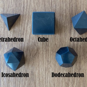 May include: Five black geometric shapes, a tetrahedron, cube, octahedron, icosahedron, and dodecahedron, are arranged on a wooden surface.