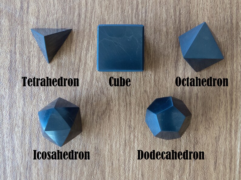 May include: Five black geometric shapes, a tetrahedron, cube, octahedron, icosahedron, and dodecahedron, are arranged on a wooden surface.