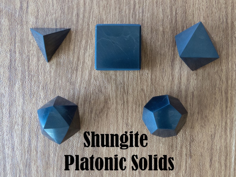 May include: Five black shungite platonic solids on a wooden surface. The solids are a cube, a tetrahedron, a dodecahedron, an octahedron, and an icosahedron. The text "Shungite Platonic Solids" is written below the solids.