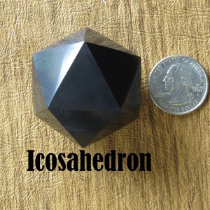May include: A black icosahedron, a 20-sided geometric shape, is shown next to a US quarter for size comparison. The icosahedron is made of a smooth, polished material.