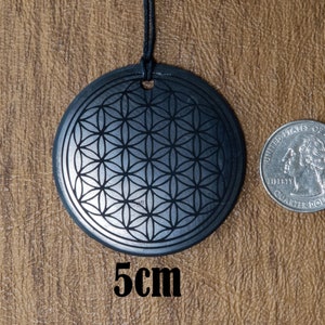 May include: A black stone pendant with a silver flower of life design. The pendant is 5 cm in diameter and is hanging from a black cord.