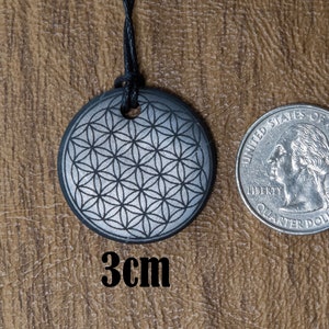 May include: A silver-colored pendant with a flower of life design, measuring 3 cm in diameter. The pendant is hanging from a black cord and is shown next to a US quarter for size comparison.