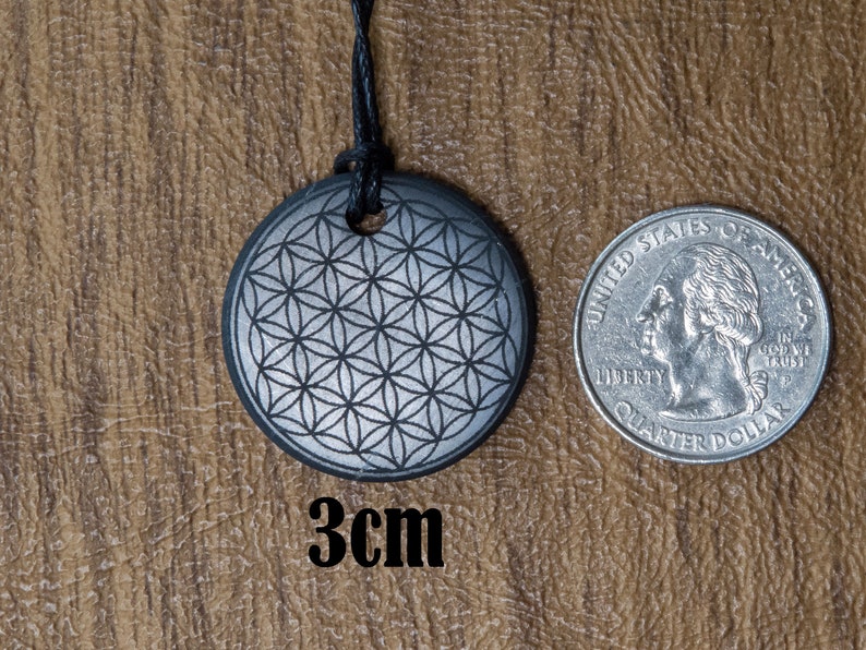 May include: A silver-colored pendant with a flower of life design, measuring 3 cm in diameter. The pendant is hanging from a black cord and is shown next to a US quarter for size comparison.