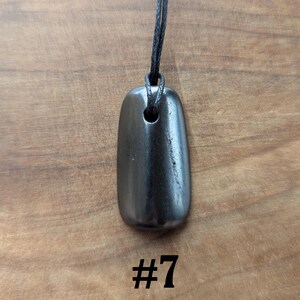 May include: A dark gray, rectangular pendant with rounded edges, suspended from a black cord. The pendant has a hole at the top for the cord. The number "#7" is printed in black below the pendant. The background is a wooden surface.