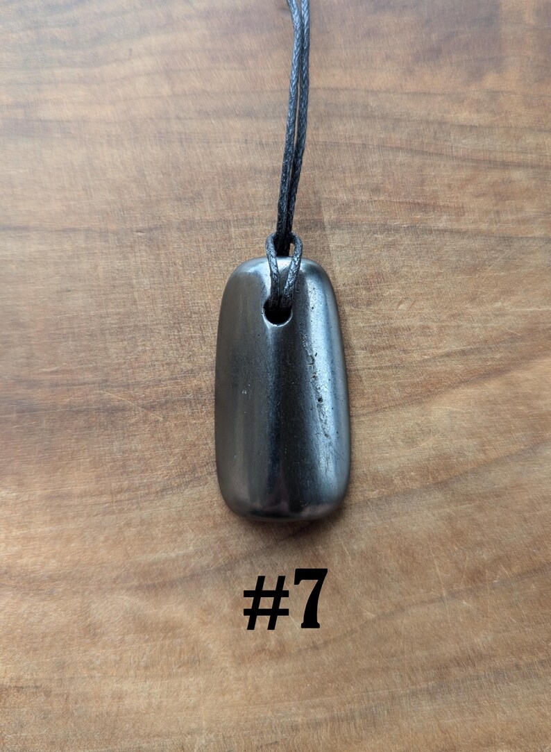 May include: A dark gray, rectangular pendant with rounded edges, suspended from a black cord. The pendant has a hole at the top for the cord. The number "#7" is printed in black below the pendant. The background is a wooden surface.