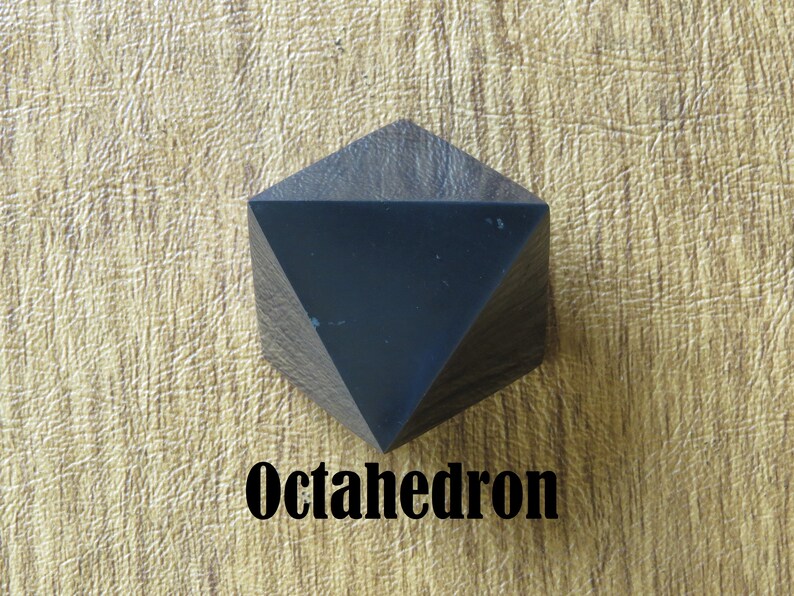 May include: A black wooden octahedron, a geometric shape with eight triangular faces. The octahedron is resting on a light brown textured surface.