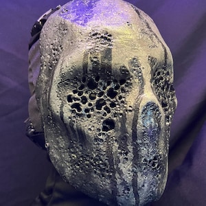 May include: A metallic mask with a textured, porous surface and dark streaks. The mask has large eye openings and a strap on the side. The color is a mix of silver, black, and hints of blue, creating a unique, industrial aesthetic.