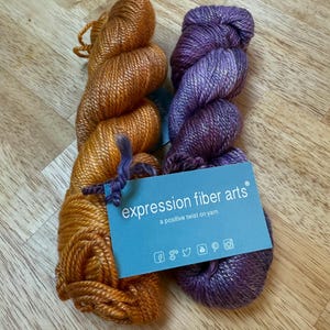 May include: Two skeins of yarn in shades of orange and purple. The orange yarn is a warm, golden hue, while the purple yarn has a deep, rich tone. A blue tag reads "expression fiber arts" with social media icons.