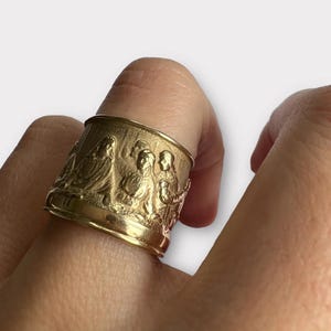 Può includere: Un anello color oro con un design a rilievo dettagliato. L'anello presenta una fascia con figure in stile classico. L'anello è indossato su un dito, mostrando la sua intricata lavorazione e il suo design.