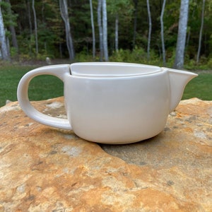 May include: A white ceramic pitcher with a handle and a spout. The pitcher is sitting on a light brown rock.