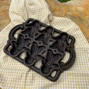 May include: A cast iron gingerbread man baking pan with four molds. The pan has two handles on the sides and is made of dark gray metal.