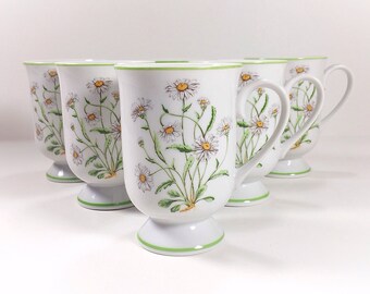 May include: Set of five white ceramic mugs with a daisy floral pattern and green trim. Each mug has a footed base.