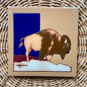 May include: A ceramic tile with a brown and tan bison on a blue and red background. The tile is signed "wtzimme".