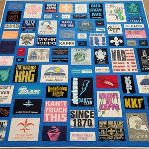 May include: A blue quilt made from various t-shirts, featuring a variety of logos, text, and graphics. The quilt includes text such as "KAPPA," "SUNNYSIDE Lake Tahoe," and "MARDI GRAS 2010." The quilt is a patchwork design.