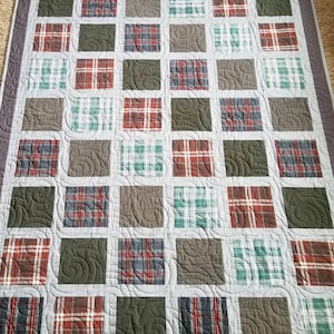 May include: A handmade quilt featuring a patchwork design with squares of plaid fabric in shades of red, green, brown, and blue. The squares are set against a light gray background with a dark gray border. The quilt is machine quilted with a swirling pattern.