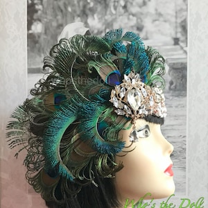 Puede incluir: Una diadema dorada con brillantes transparentes y un colgante de cristal, adornada con un gran tocado de plumas de pavo real. El tocado está hecho de plumas de pavo real verdes y azules.