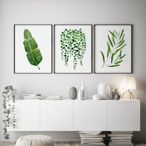 May include: Three framed watercolour prints of green foliage. The prints are hung on a white wall above a white cabinet with a white countertop. The cabinet is decorated with vases and other decorative items.