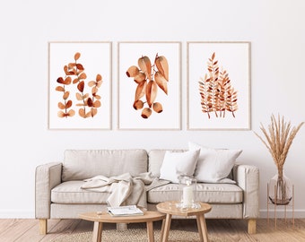 May include: Three framed watercolour prints of brown leafy branches on a white background. The prints are hanging on a white wall above a light brown sofa with white throw cushions.