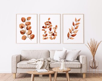 May include: Three watercolor prints of brown eucalyptus leaves on a white background. The prints are framed in a simple wooden frame and are hung on a white wall above a beige sofa.