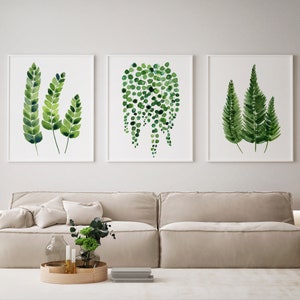 Op de afbeelding: Drie ingelijste aquarelprints van groen gebladerte. De prints hangen aan een witte muur boven een lichtbeige bank.