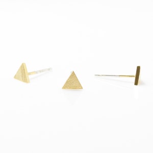 Puede incluir: Un par de pendientes de triángulo de oro con acabado cepillado. Los pendientes están sobre un fondo blanco.