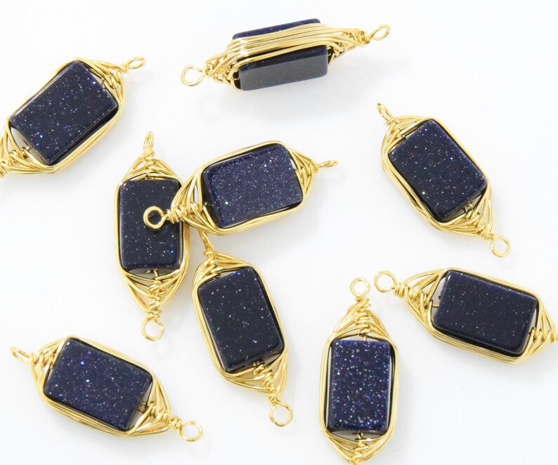 May include: Gold-colored wire-wrapped pendants with rectangular, dark blue, glittery stones. The pendants are arranged on a white surface, showcasing their intricate wirework and the sparkling stones.