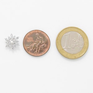 May include: A silver snowflake charm with clear rhinestones, next to a US penny and a Euro coin for size comparison.