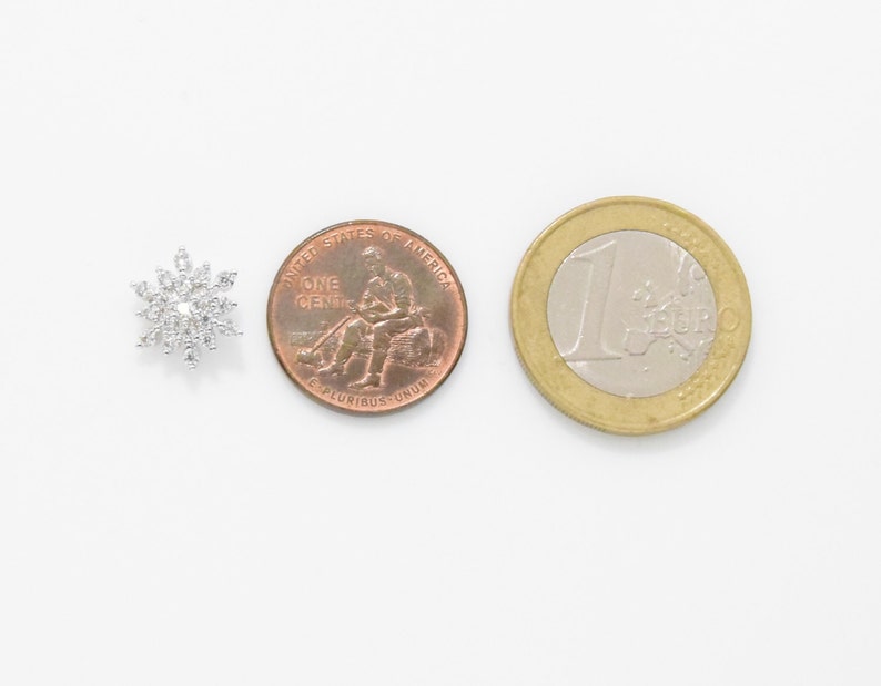 May include: A silver snowflake charm with clear rhinestones, next to a US penny and a Euro coin for size comparison.