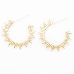 Op de afbeelding: Een paar gouden creolen met een zonnestralenontwerp. De oorbellen zijn gemaakt van metaal en hebben een puntige, stekelige rand.