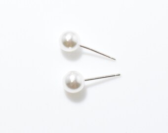 Op de afbeelding: Twee paar witte parelstud-oorbellen met zilveren stiftjes worden getoond tegen een witte achtergrond. De parels hebben een gladde, ronde vorm en de stiftjes zijn dun en metaalachtig. De oorbellen zijn eenvoudig en elegant.