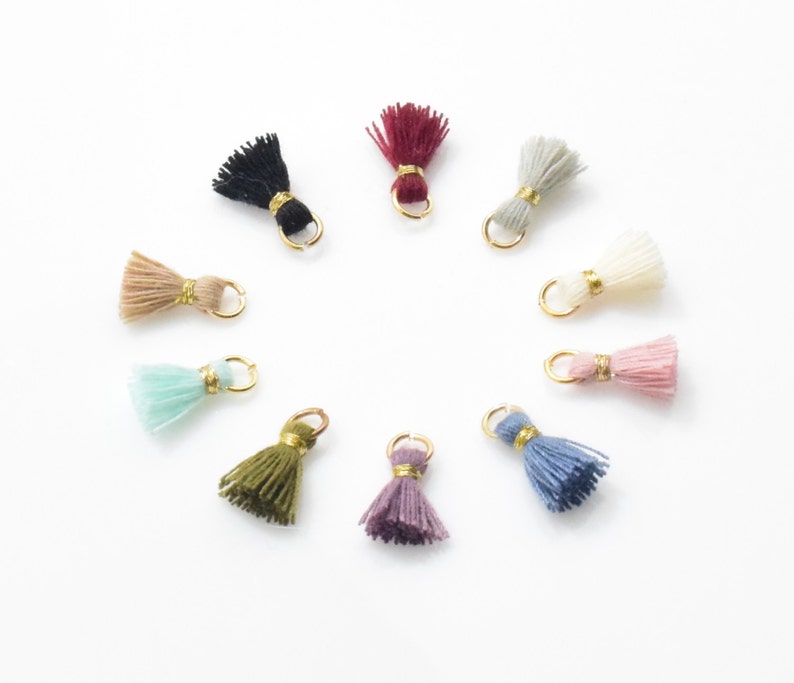 May include: Assortment of ten colorful tassel charms with gold jump rings. The tassels are made of various colors including black, white, pink, blue, green, beige, and purple.